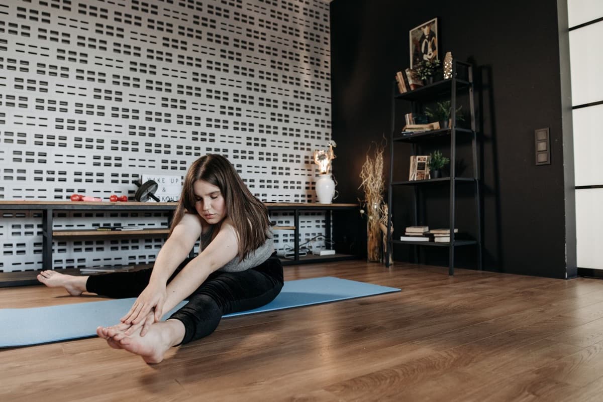 A woman sitting on a yoga mat at home doing a simple seated forward-fold hamstring stretch