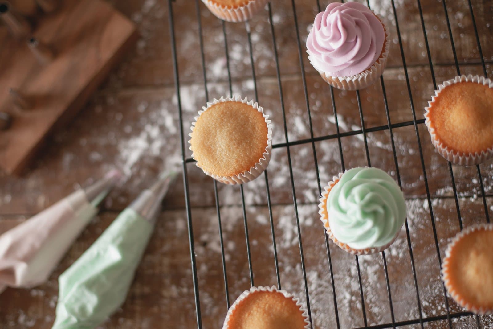 Unfrosted and freshly piped cupcakes cooling on a wire rack next to pastel piping bags