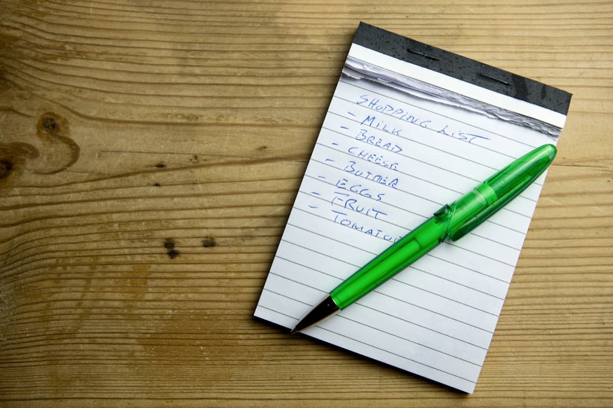 A handwritten shopping list (milk, bread, cheese, butter, eggs, fruit, tomatoes) on a notepad with a green pen, on a wood table