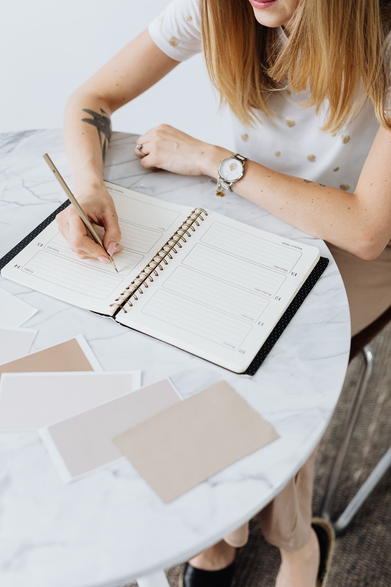 A woman writing in a weekly planner at a marble desk, with color swatches scattered nearby