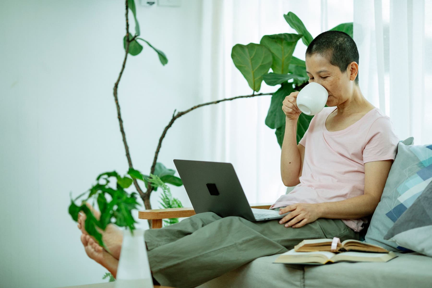 A woman with a short buzz cut sitting on a sofa in a bright, plant-filled living room, drinking from a white mug with her eyes closed, a laptop and open book on her lap