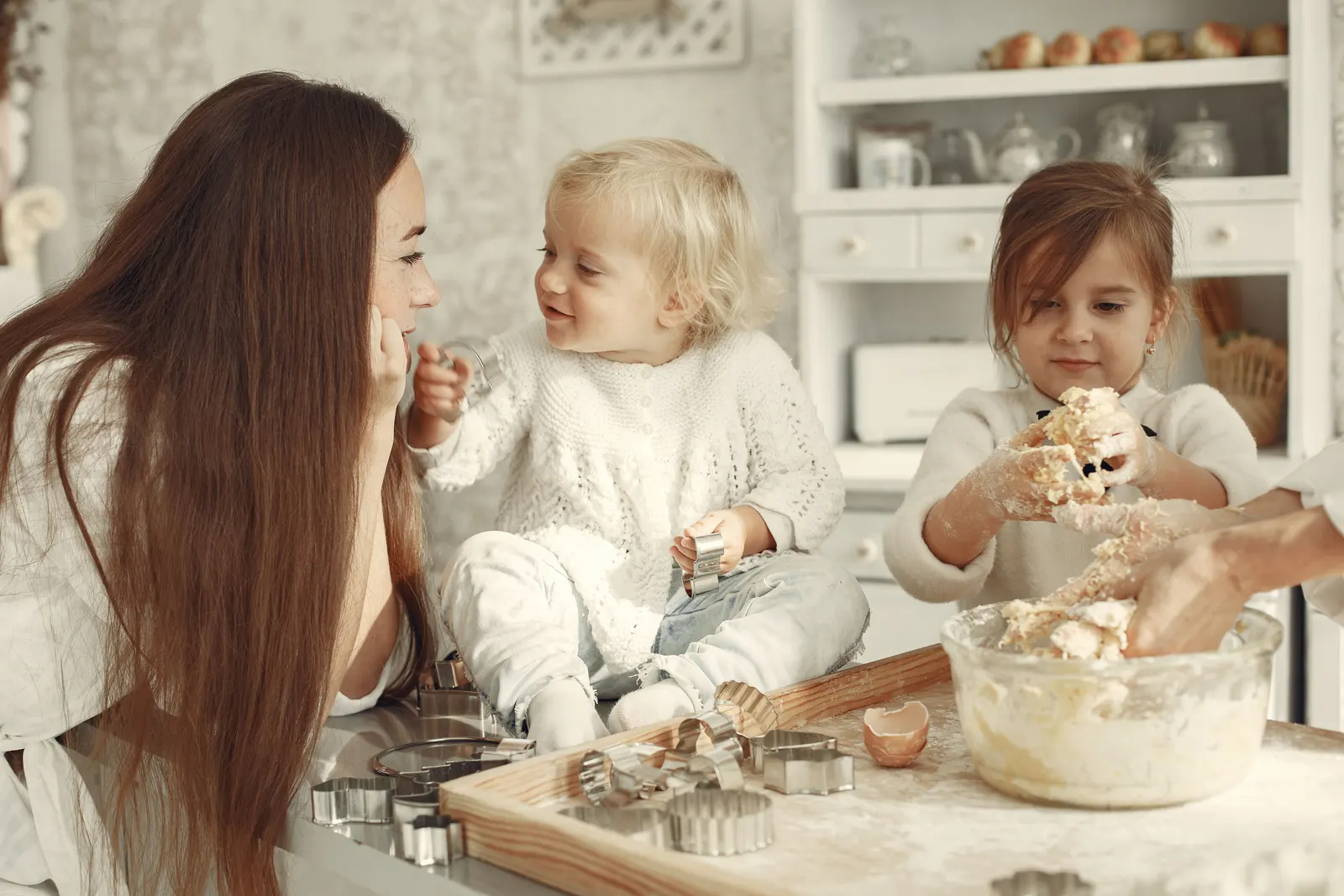 A mother and her two young daughters mixing cake batter together in a bright home kitchen, surrounded by cookie cutters and a mixing bowl