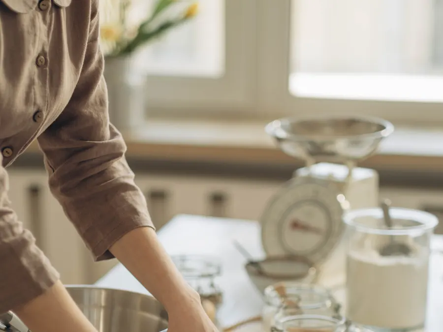 A home baker reading a printed recipe book while gathering flour, a mixing bowl, and a kitchen scale on the counter