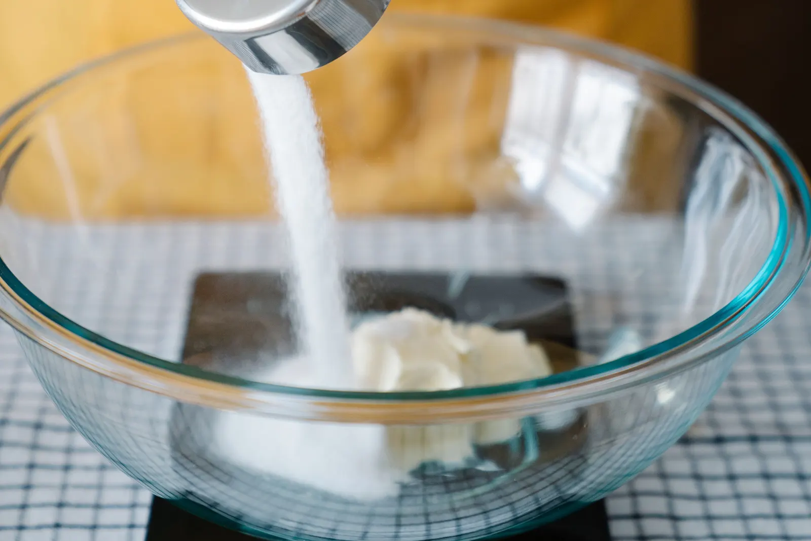A hand pouring sugar from a measuring cup into a mixing bowl on a kitchen counter