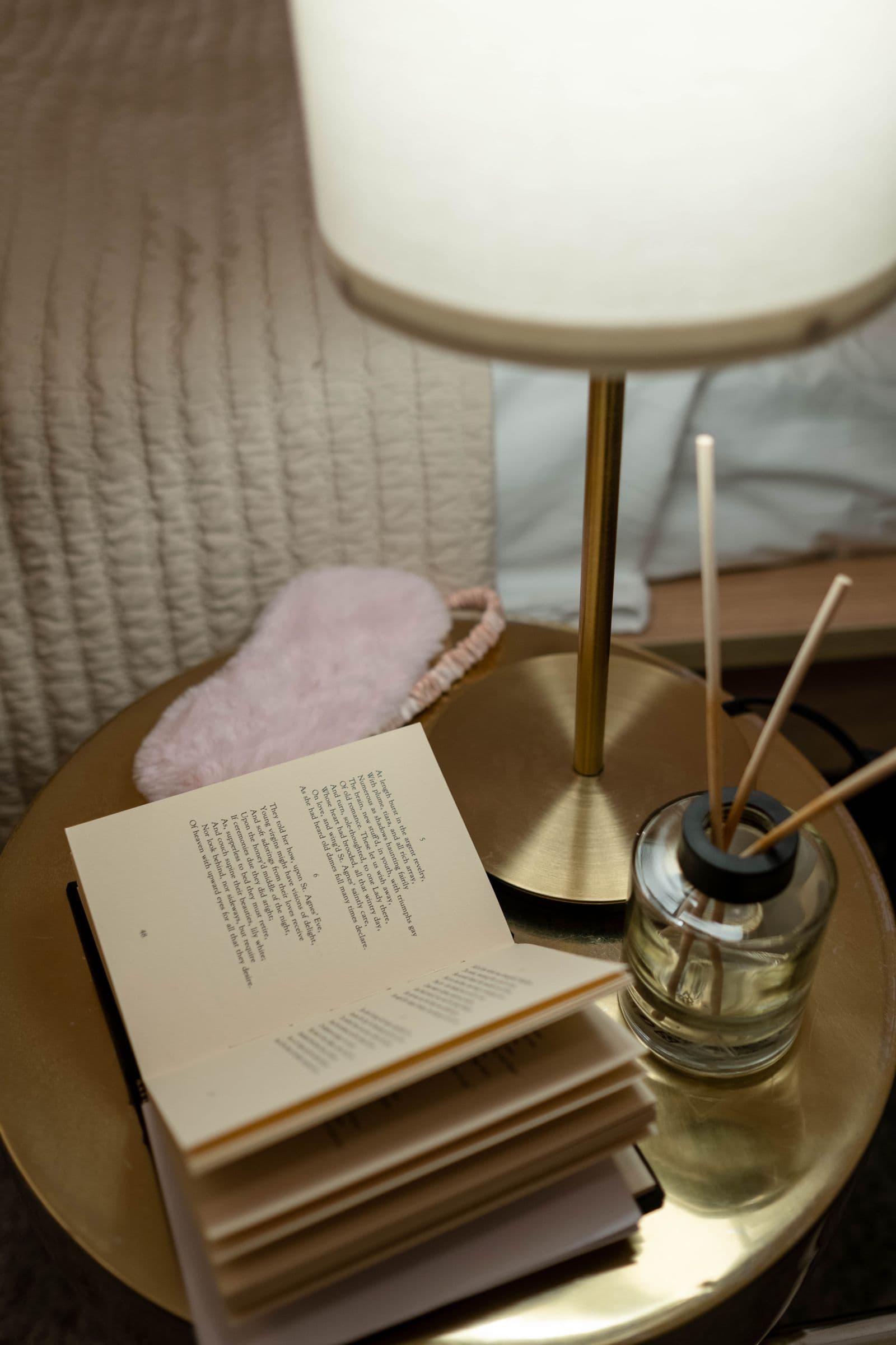A softly lit bedside table with a lamp, an open book, a sleep mask, and a reed diffuser