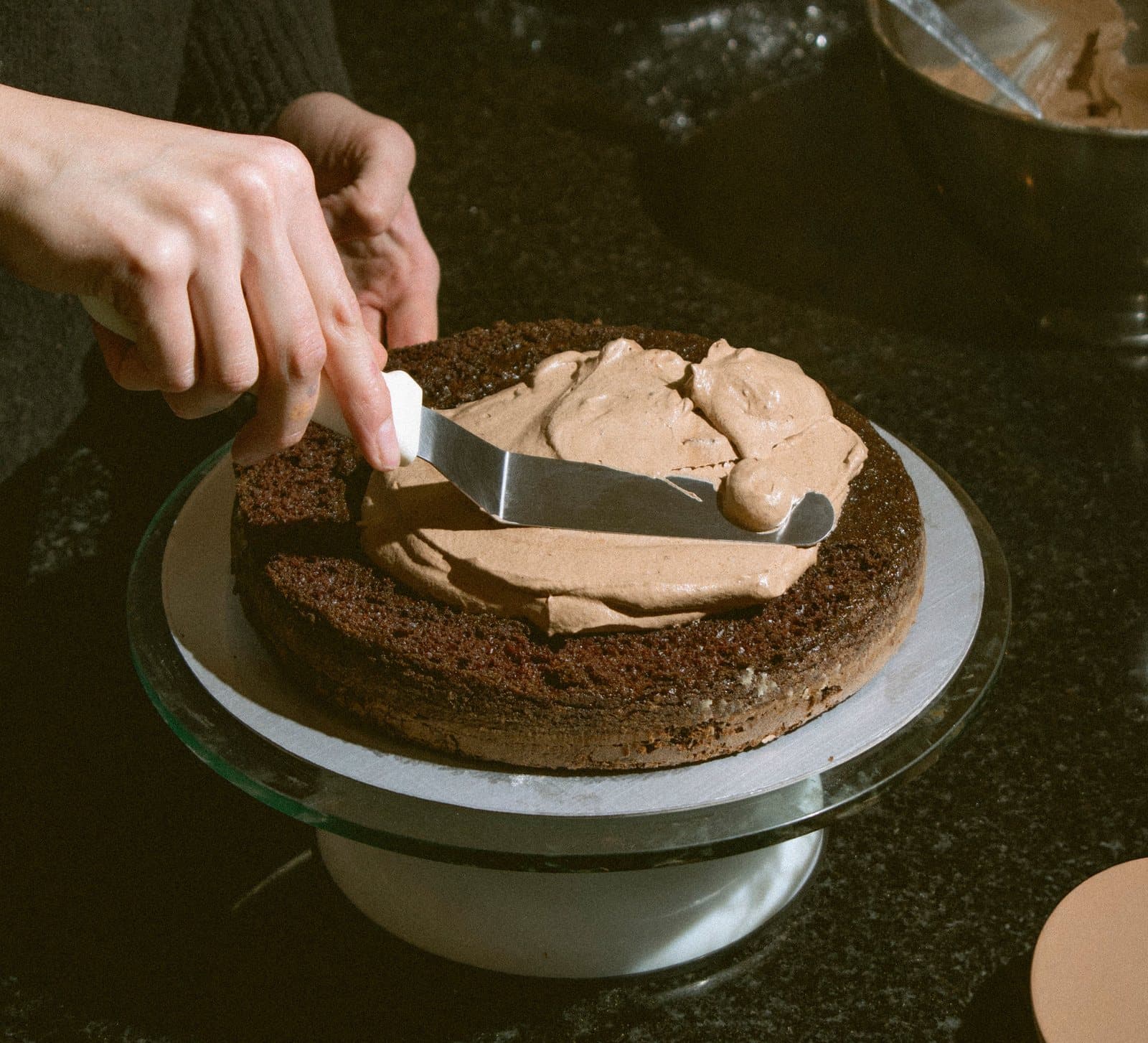 A hand smoothing chocolate frosting onto a cake on a turntable with an offset spatula