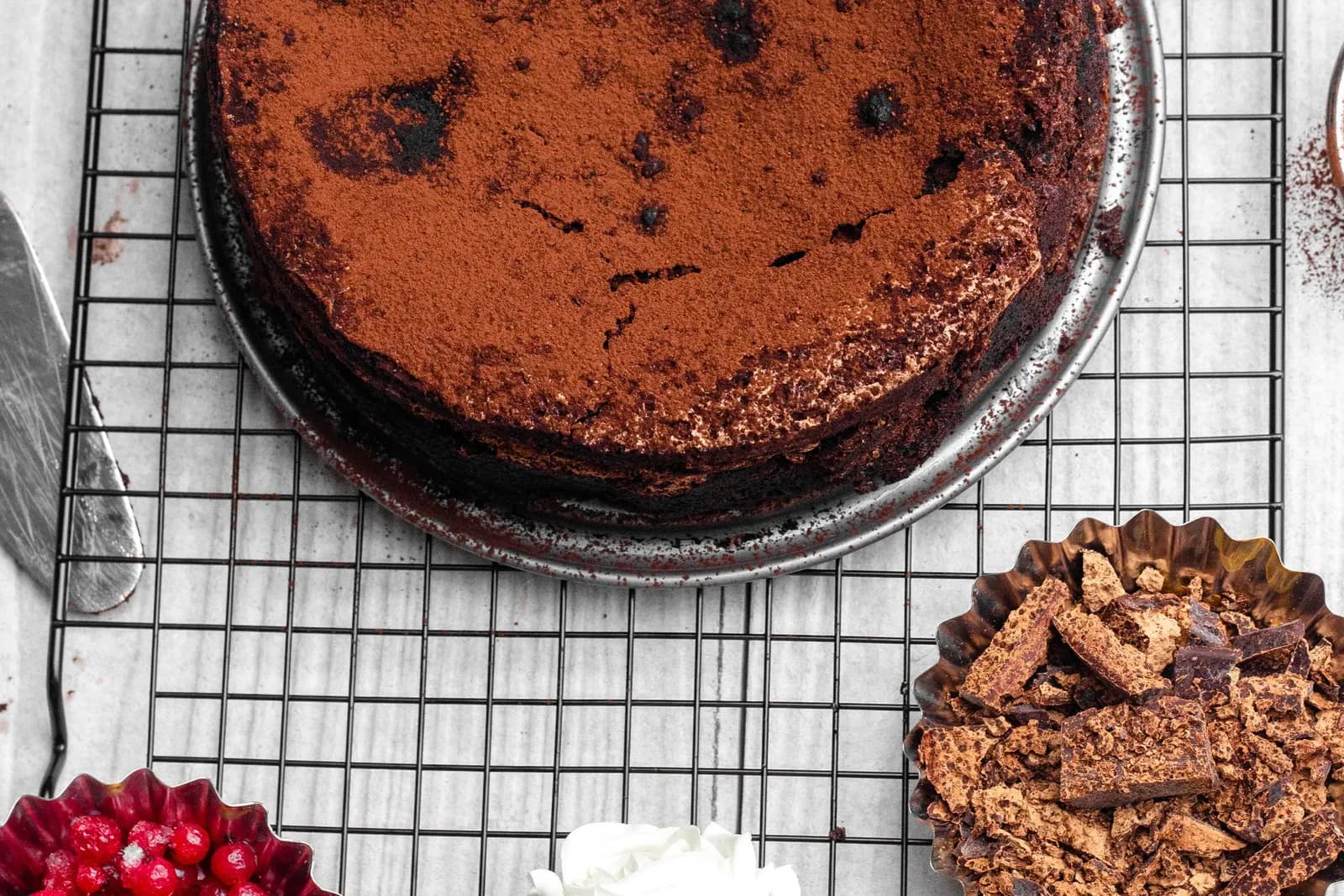 A chocolate cake dusted with cocoa powder, showing a cracked top, cooling on a wire rack