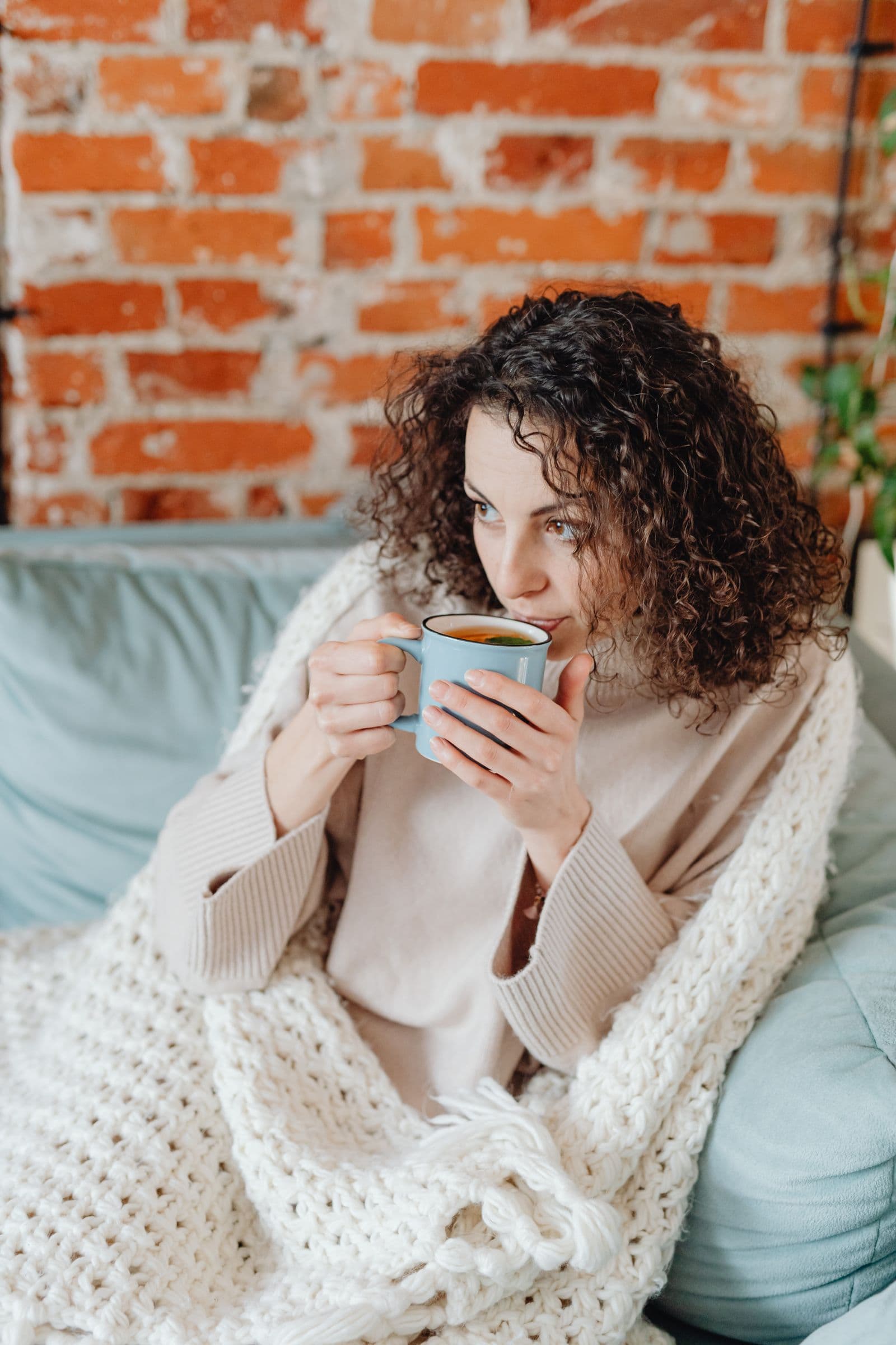 A woman wrapped in a chunky knit blanket on a sofa, sipping from a mug of tea in a cozy room with an exposed brick wall