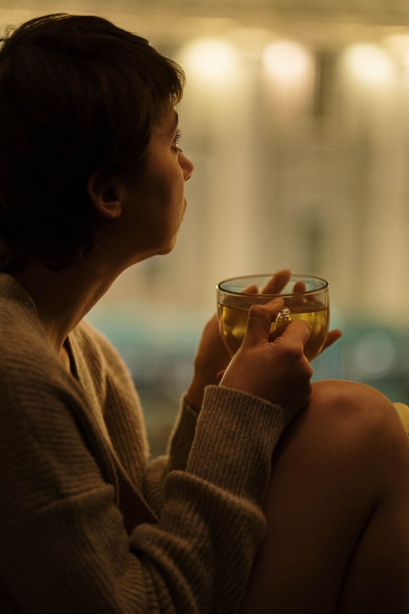 A woman in silhouette holding a cup of tea and looking thoughtfully out a window in warm evening light