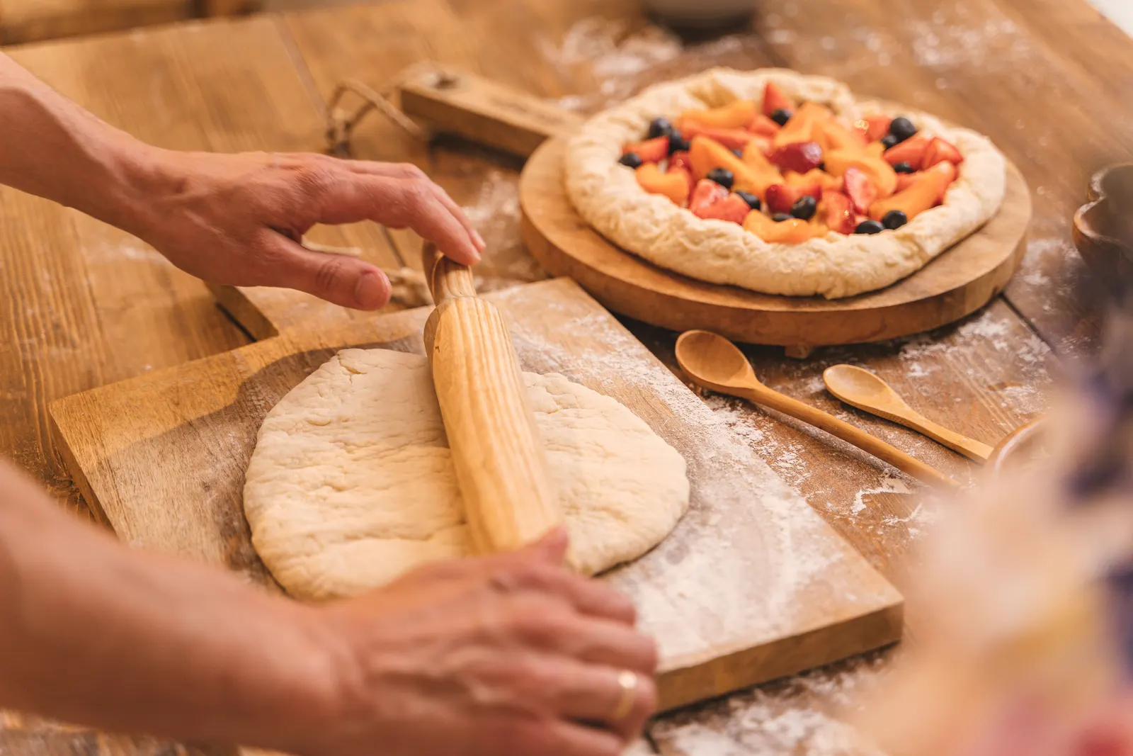 Hands rolling out dough next to a fruit pie on a wooden table
