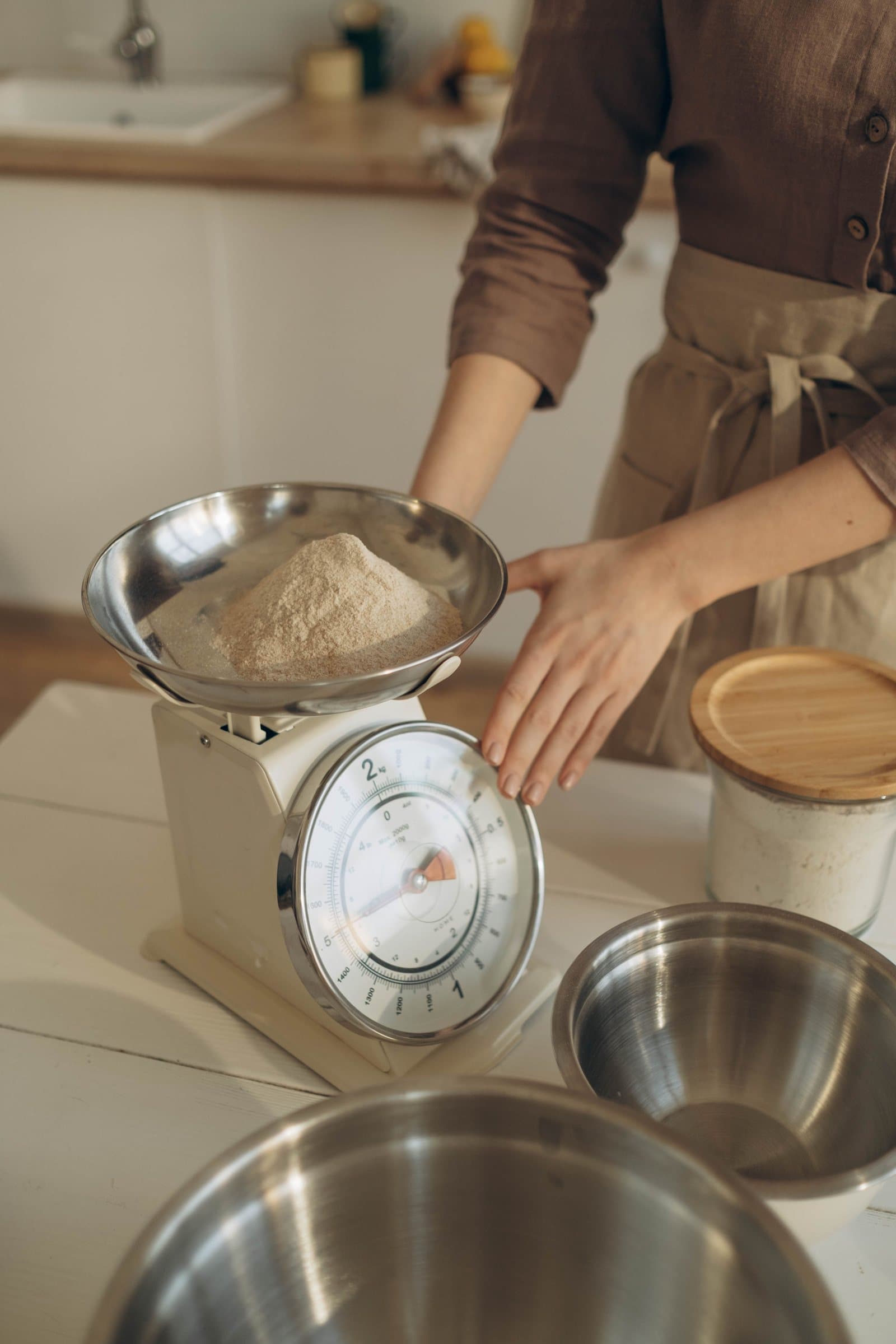 A woman weighing flour on an analog kitchen scale, with mixing bowls nearby
