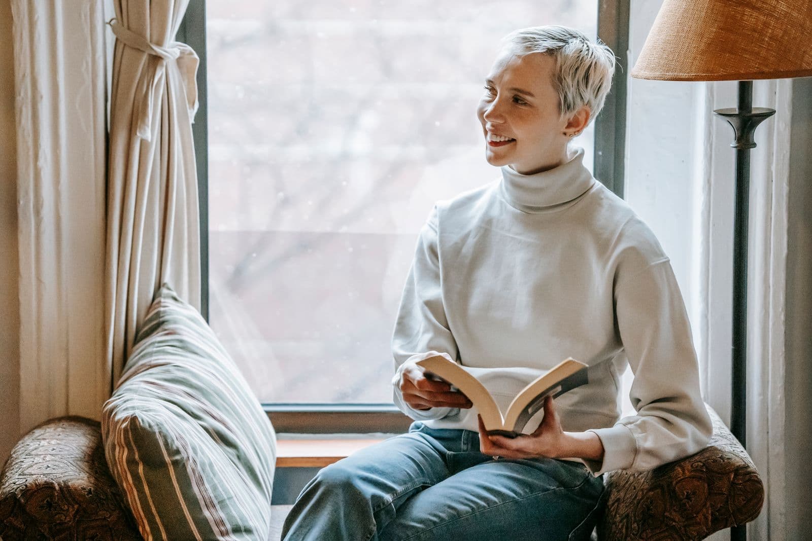 A smiling woman reading a book in a cozy window seat, soft daylight coming through the window