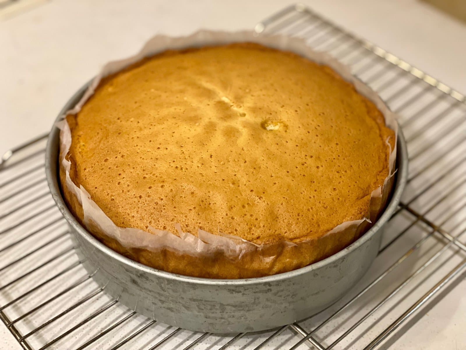 A baked round sponge cake cooling in its pan on a wire rack, with a faint crack across the domed top