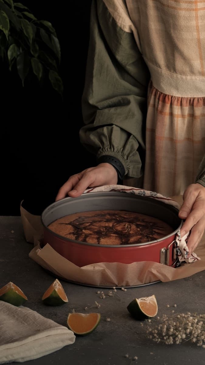 A person lifting the springform ring off a freshly baked cake, with citrus wedges and linen on the table